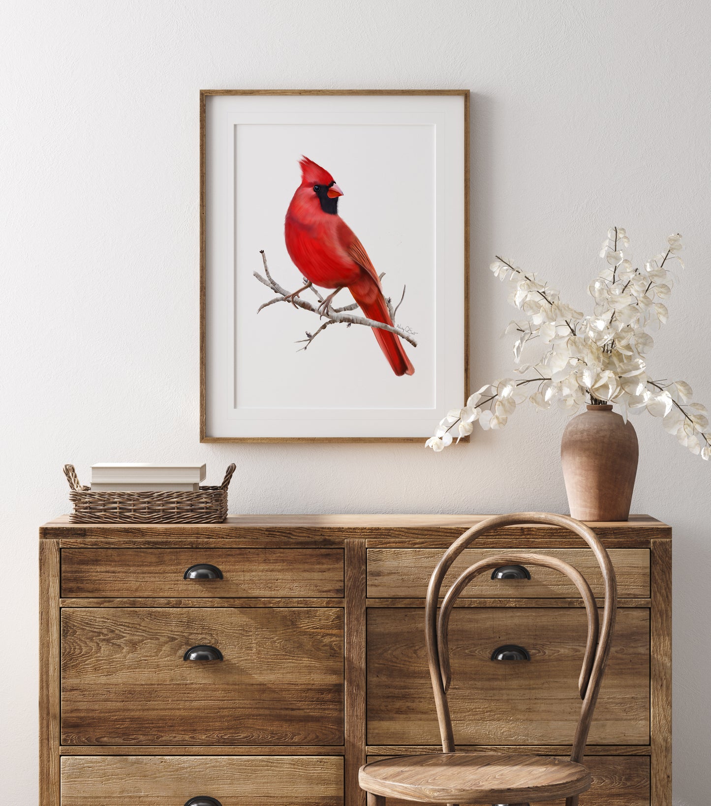 Illustration of a red cardinal bird sitting on a brow twig branch without leaves. The background of the artwork is plain white. The print is shown matted in a wooden frame and mounted on a wall above a wooden chest of drawers. Art by Nicky Quartermaine Scott