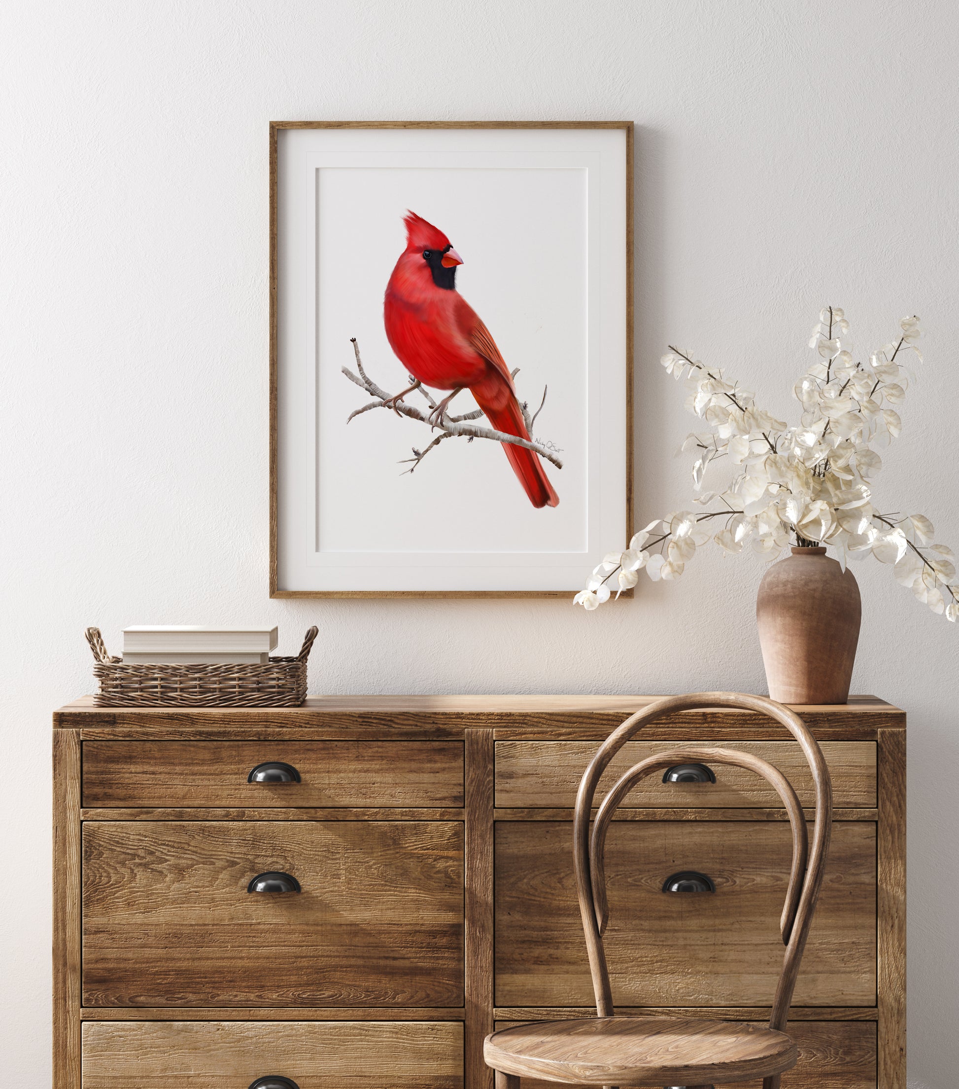 Illustration of a red cardinal bird sitting on a brow twig branch without leaves. The background of the artwork is plain white. The print is shown matted in a wooden frame and mounted on a wall above a wooden chest of drawers. Art by Nicky Quartermaine Scott