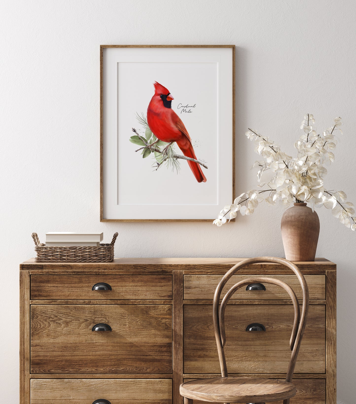 Framed picture of a red cardinal bird in leaves mounted against a while wall and hung above wooden dresser with a vase and basket.