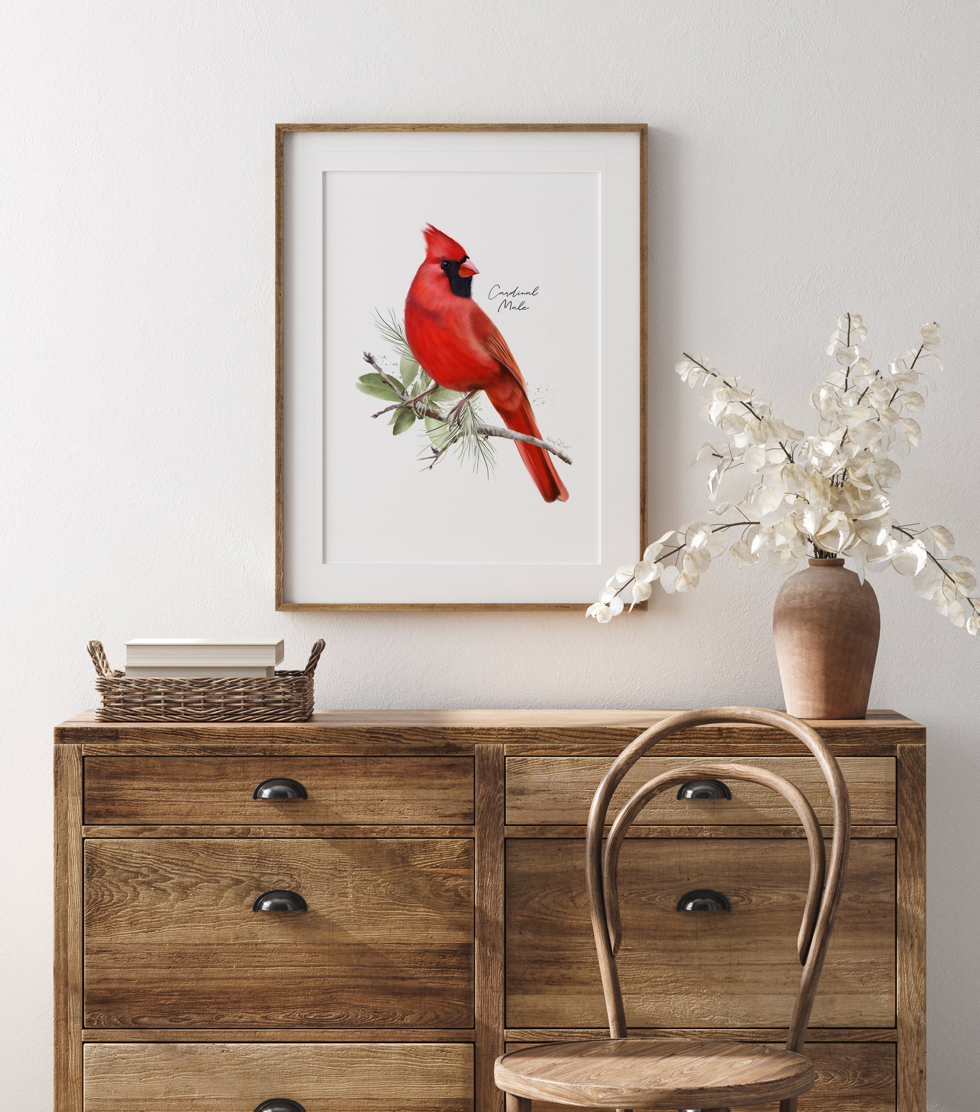Framed picture of a red cardinal bird in leaves mounted against a while wall and hung above wooden dresser with a vase and basket.
